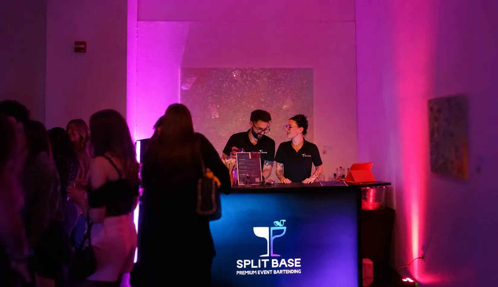 Two Split Base bartenders serving a party behind a neon-lit bar
