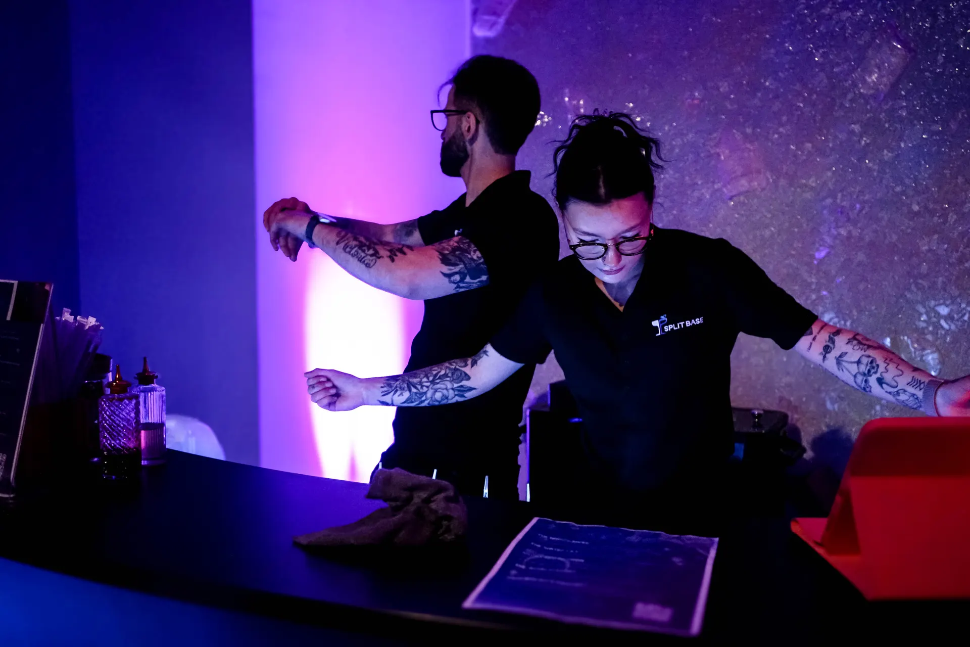 Two Split Base bartenders working a neon-backlit bar in a dark room
